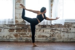 woman in black leggings and tank top doing dancer post in front of a rustic brown and white wall